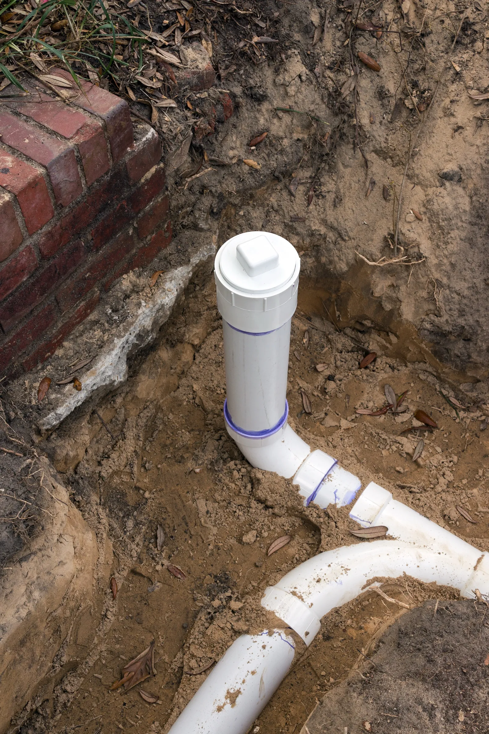 Capped two-way sewer cleanout uncovered in a trench beside a brick home foundation, the access point for a hydrostatic test.
